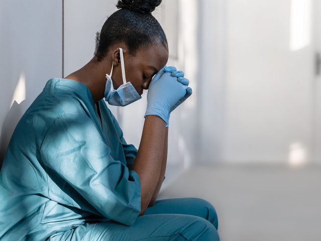 Tired depressed female african scrub nurse wears face mask blue uniform gloves sits on hospital floor. Exhausted sad black doctor feels burnout stress of corona virus frontline protection pray at work